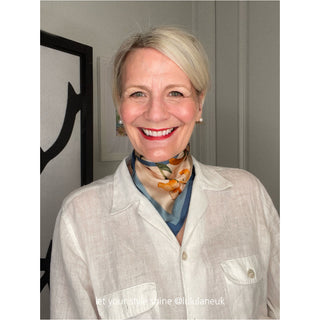A stylish blonde woman in a white blouse wears an elegant Iris print silk bandana scarf with orange flowers and butterfly design on cream background with blue border, paired with elegant pearl stud earrings.