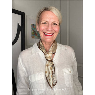 A stylish blonde woman in a white blouse wears a beige and brown silk scarf in earthy tones, featuring a bohemian paisley and polka dot print with contrasting brown edges, paired with elegant pearl stud earrings.