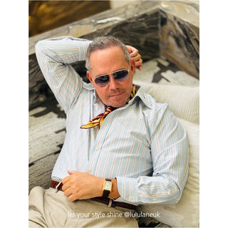 Stylish man wearing a silk neckerchief in red, black and beige yellow tones with a light blue striped shirt, sunglasses, and wristwatch while relaxing on a sofa