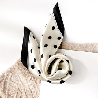 Elegant white silk scarf with black polka dots and black border, loosely draped over a beige knit sweater, showcasing its lustrous texture