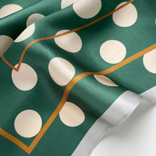 Jade green silk scarf with cream polka dots and gold border, showing luxurious fabric texture with gentle folds and white edge trim.