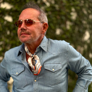 Styling example: Baroque silk neckerchief with blue and orange pattern worn with light denim shirt and amber sunglasses in outdoor setting