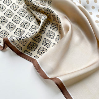 Beige silk scarf with intricate black paisley pattern on one side, solid cream color on the other, featuring elegant brown border trim