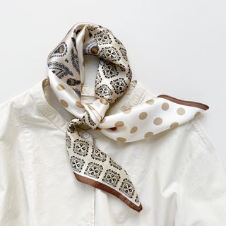 Beige silk bandana scarf with paisley patterns and polka dots, elegantly knotted around white shirt collar with brown trim detail