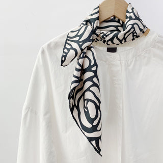 a cream white base women's silk scarf with charcoal black abstract rose print and hand-rolled edges, knotted as a neck scarf, paired with a women's white turtleneck shirt