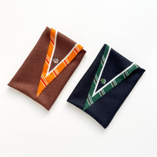 Two folded diamond-shaped scarves: brown with orange trim and black with green trim, both featuring English plaid pattern and small logo embellishment.