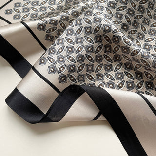 Luxurious silk bandana scarf with detailed geometric pattern in black and beige, featuring elegant black border stripes and intricate eye motifs