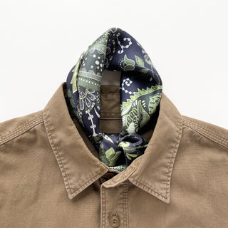 Forest green paisley silk scarf tied at neck of tan button-up shirt, showcasing intricate navy and green pattern with hand-rolled edges