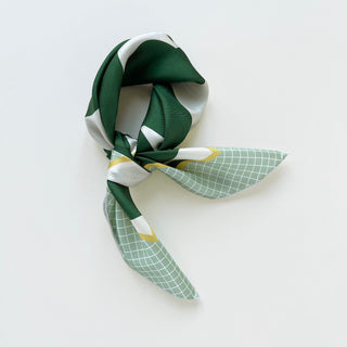 Small silk scarf featuring geometric pattern with deep green blocks, white accents, and mint green grid design with yellow dots, elegantly folded
