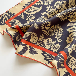 Luxurious navy and beige silk scarf with intricate paisley pattern, featuring hand-rolled orange border and elegant sheen on twill fabric.