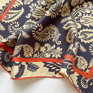 Luxury silk scarf with navy and beige paisley pattern, featuring vibrant orange border and hand-rolled edges, shown draped to display its intricate design
