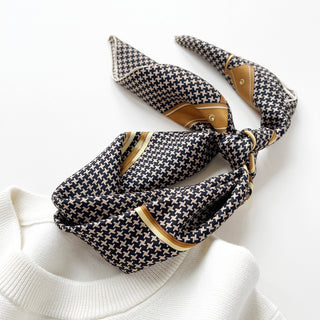 Black and beige houndstooth silk scarf tied elegantly with golden accents, displayed on white fabric showing its luxurious texture and classic pattern.
