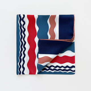 Square silk scarf with hand-rolled edges featuring wave pattern in navy blue, teal, red, and terracotta on white background, partially folded