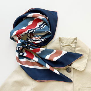 Silk scarf with wave pattern in navy blue, red, and earthy tones, knotted elegantly on beige jacket collar showing hand-rolled edges