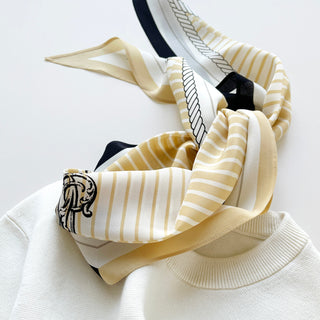 Long silk scarf in pale yellow and beige stripes with black accents, decorative monogram, and rope border design, draped on cream sweater