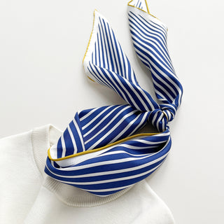 Triangle silk scarf with navy blue and white stripes, gold-trimmed edges, elegantly tied and draped on a cream sweater collar