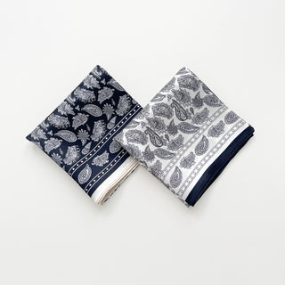 Two folded silk bandana scarves with paisley pattern - one navy blue with white motifs and one white with navy patterns, both featuring elegant border details