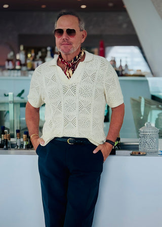 Stylish man wearing cream white knitted polo shirt with red, black and beige geometric silk neckerchief, navy blue trousers, and sunglasses for a smart casual look in a bar setting