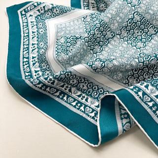 Teal luxury silk scarf with intricate white geometric patterns and decorative bird border, featuring hand-rolled edges and shimmering twill texture