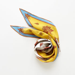Diamond-shaped silk scarf in yellow and ivory with floral print, displaying in an elegant swirl on a white background, showcasing blue and rust border accents with orange hand-rolled edges