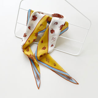 Diamond-shaped silk scarf in yellow and ivory with floral print, hanging on a white hanger on a white background, showcasing blue and rust border accents with orange hand-rolled edges.