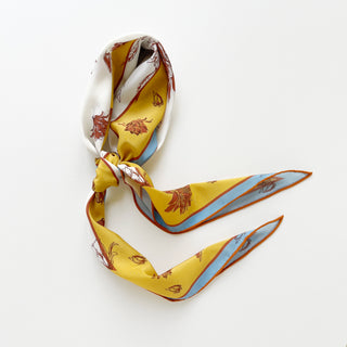 Diamond-shaped silk scarf in yellow and ivory with floral print, tied in a loose knot on a white background, showcasing blue and rust border accents with orange hand-rolled edges.