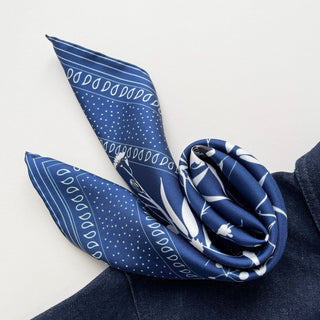 Blue silk neckerchief with white botanical pattern, featuring dots, leaf motifs and stripes, elegantly styled on denim fabric showing luxurious sheen and texture.