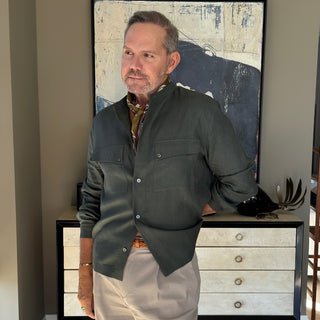 Men’s silk and cotton neck scarf in olive with stylised bird print, styled with a dark green jacket, pleated trousers.