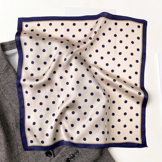 Elegant silk bandana scarf with champagne center featuring navy blue polka dots and navy border, displayed on a patterned surface showing its luxurious texture