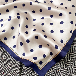 Elegant silk bandana scarf with navy blue polka dots on cream background, finished with navy trim and gold border, displayed on textured fabric.