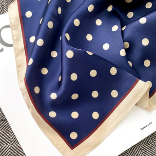 Navy blue silk scarf with ivory polka dots, featuring champagne border trim with burgundy accent, displayed in elegant folds showing luxurious fabric texture.