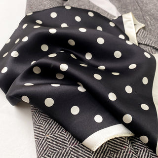 Navy blue silk bandana scarf with white polka dots and cream border, elegantly draped over patterned fabric showing luxurious sheen