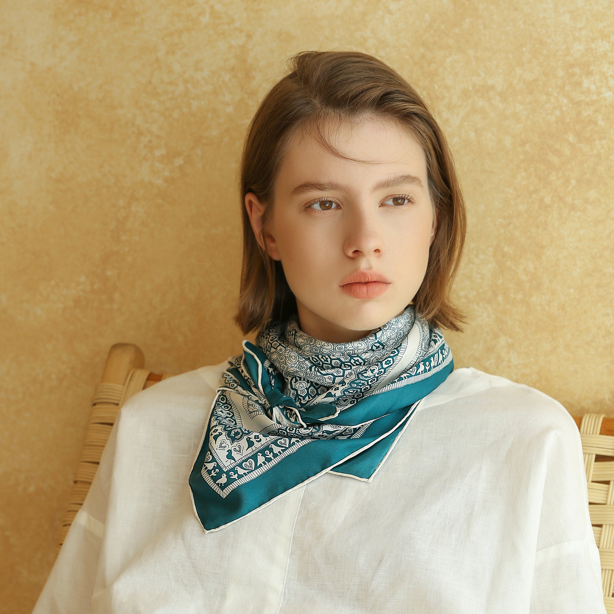 a turquoise blue square silk scarf with birds, flowers and trees prints worn by a young lady as a neck scarf