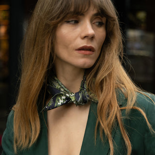 Woman wearing a navy and green patterned silk bandana scarf by LuluLane in a green blazer holding a red handbag on a street.
