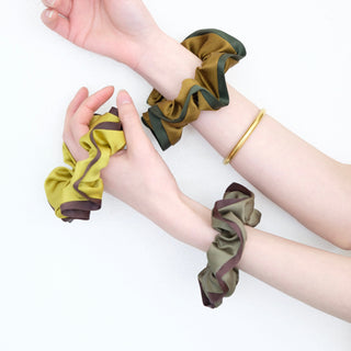 Three luxurious silk scrunchies displayed on wrists in earth tones - mustard with brown trim, olive with burgundy edge, and khaki with green border.