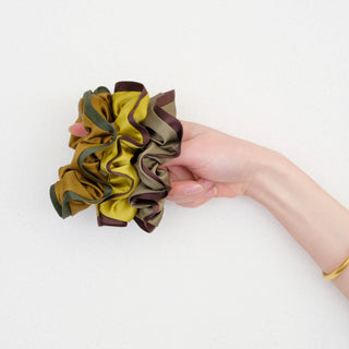Hand holding three ruffled silk scrunchies in earth tones of gold, olive, and khaki with contrasting edges, showcasing their luxurious texture and volume