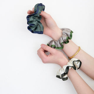 Hands displaying three silk scrunchies with contrast edges - green with navy, silver with green, and cream with black - against white background