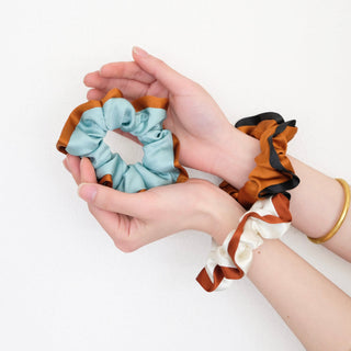 Hands holding luxury silk scrunchies in blue with copper trim, black with copper trim, and ivory with copper trim, showcasing contrast edge design