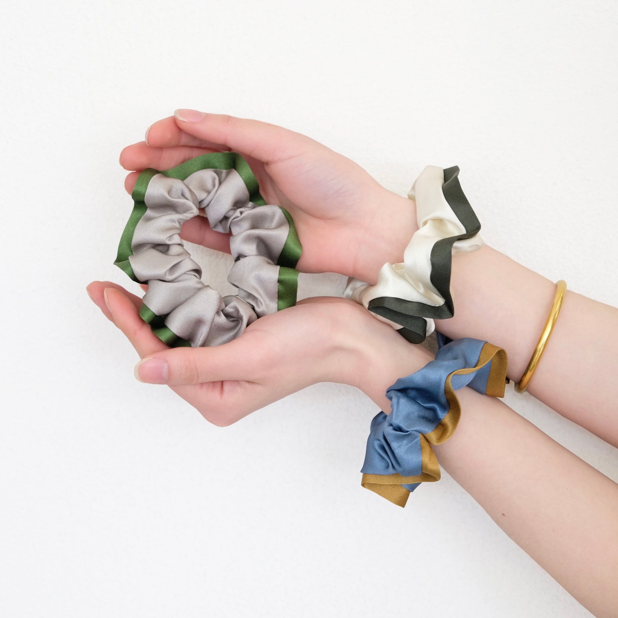 three silk scrunchies in different colours holding by a pair of woman's hands, one is khaki with green edge, one is creamy white with dark green edge, one is Egyptian blue with bronze edge 
