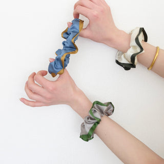 Three silk scrunchies worn on wrists - blue with copper edge, cream with black trim, and khaki with green accent - showcasing luxurious ruffled texture