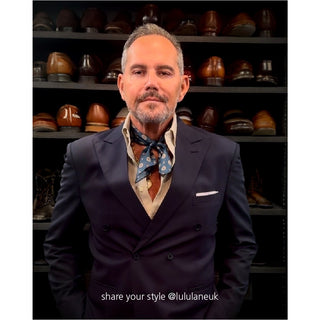 Denim blue silk neckerchief with medallion pattern styled with navy blazer and white shirt, displayed against backdrop of luxury footwear