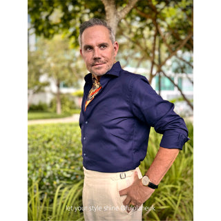 Man wearing a navy blue shirt with rolled-up sleeves and a luxury silk scarf with navy and beige paisley pattern featuring vibrant orange border, standing outdoors. 
