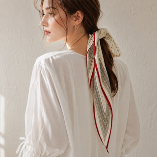 A woman shown from the back wearing a white blouse and a patterned silk scarf tied around her hair, set against a textured off-white wall.