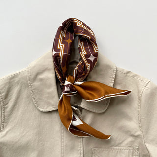 Vintage 70s silk neckerchief in burgundy and amber with geometric pattern, styled on beige jacket collar showing luxurious hand-rolled edges