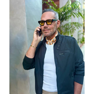 Man wearing unisex chess board pattern silk square scarf in golden squash yellow and navy by LuluLane in dark blue racer jacket, talking on a phone with plants in the background.