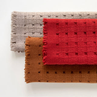Three pure cashmere scarves featuring openwork jacquard design in red, mustard brown, and beige on a light grey background.