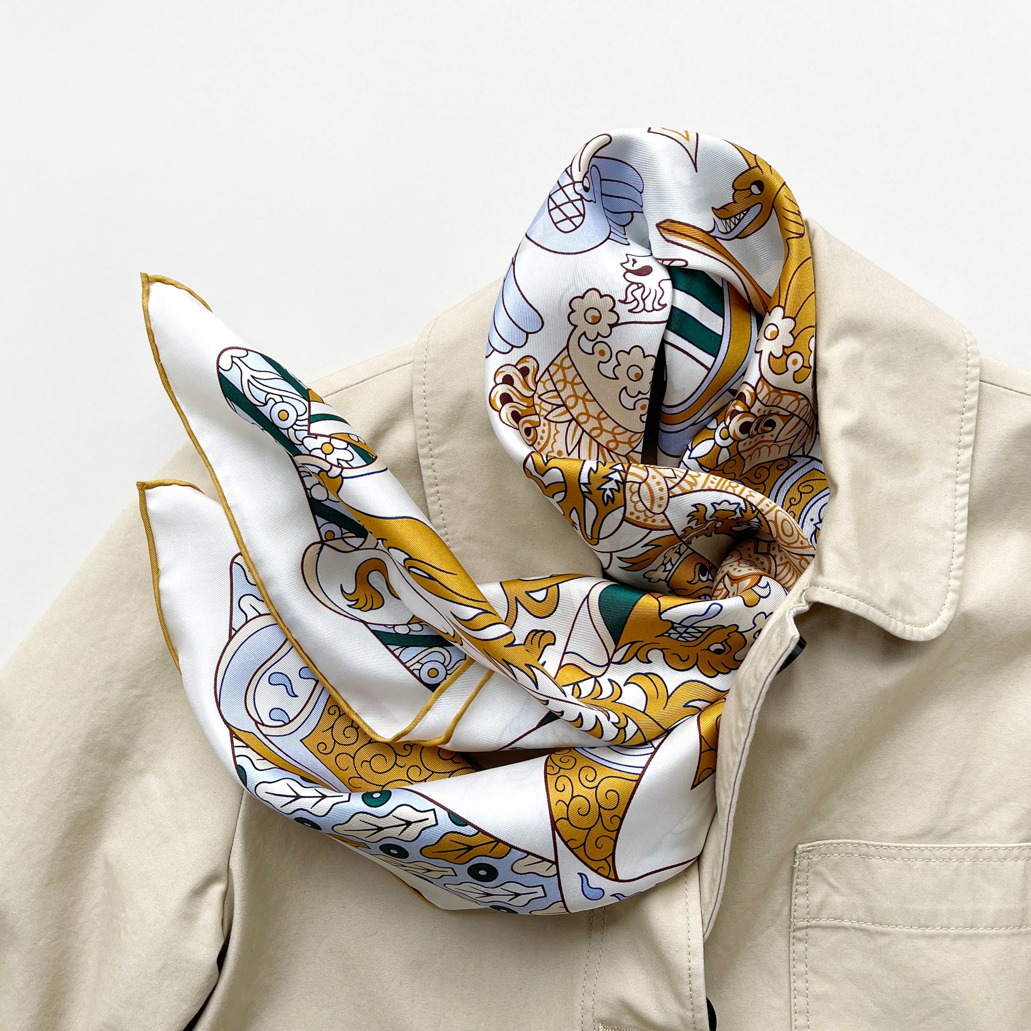 a large square silk scarf featuring Greek mythology print in mustard yellow, green and sky blue on a white base, with mustard yellow hand-rolled edge, tied as a neck scarf, paired with a beige coat