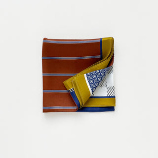 Folded navy silk square neckerchief with geometric patterns featuring blue, rust, gold and white sections with detailed diamond motifs and dotted designs