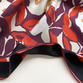 Close-up of a silk bandana scarf by LuluLane with burgundy background, cream white floral print, and orangey red leaves, framed by black border with hand-rolled edges.
