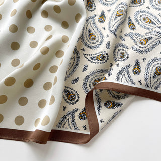 Close-up of Beige silk scarf by LuluLane with intricate black paisley pattern on one side, solid cream colour on the other, featuring elegant brown border trim